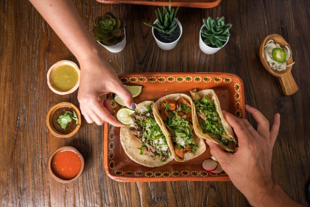 Hands reaching for tacos with various toppings on a decorated ceramic plate, accompanied by three small bowls of different sauces, set on a wooden table with small potted plants.