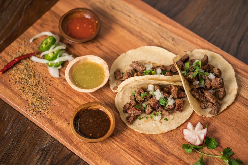 Two tacos filled with seasoned meat, garnished with cilantro and onions, served on a wooden board with three small bowls of different sauces, and additional garnishes including sliced jalapeños and radishes.